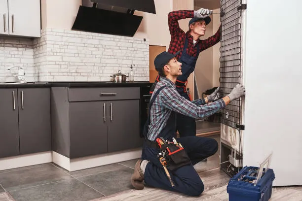 A technician performing maintenance on a stainless steel refrigerator using a specialized vacuum tool on a wooden floor.