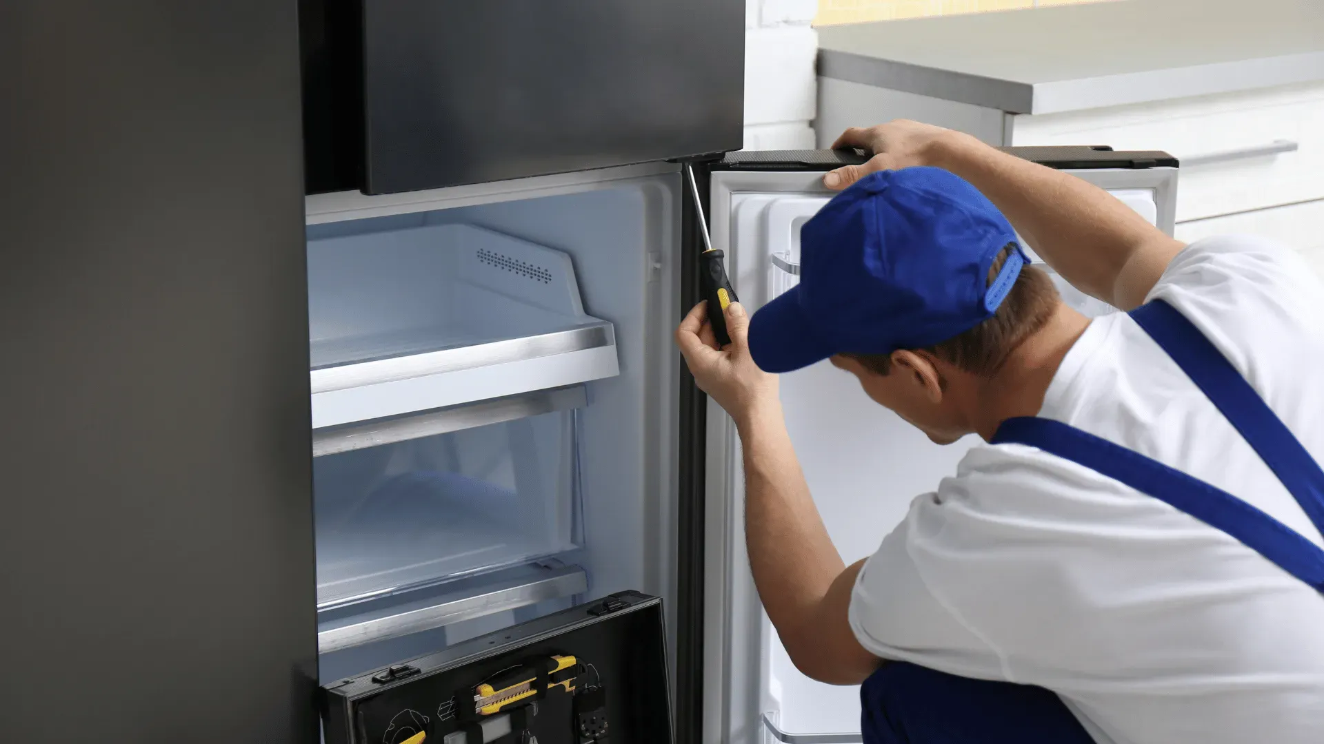 Professional 24x7 Service UAE technician in a blue uniform repairing the internal thermostat and light assembly of a silver refrigerator.